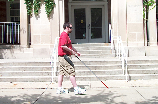 A student with a white cane walking outside Fell Hall.
