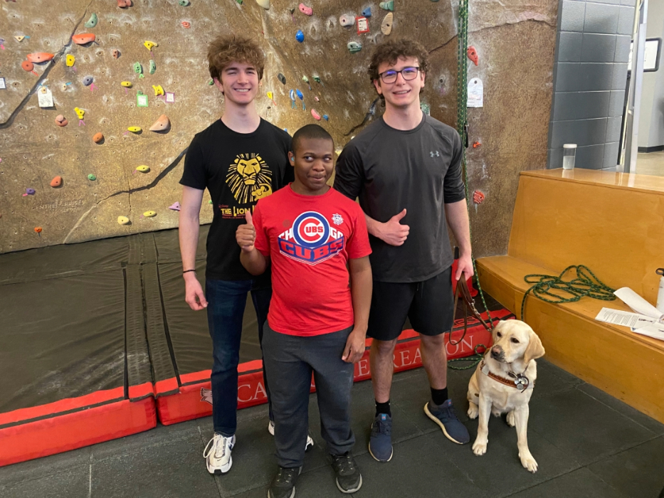 Three students and one guide dog standing in front of a climbing wall.